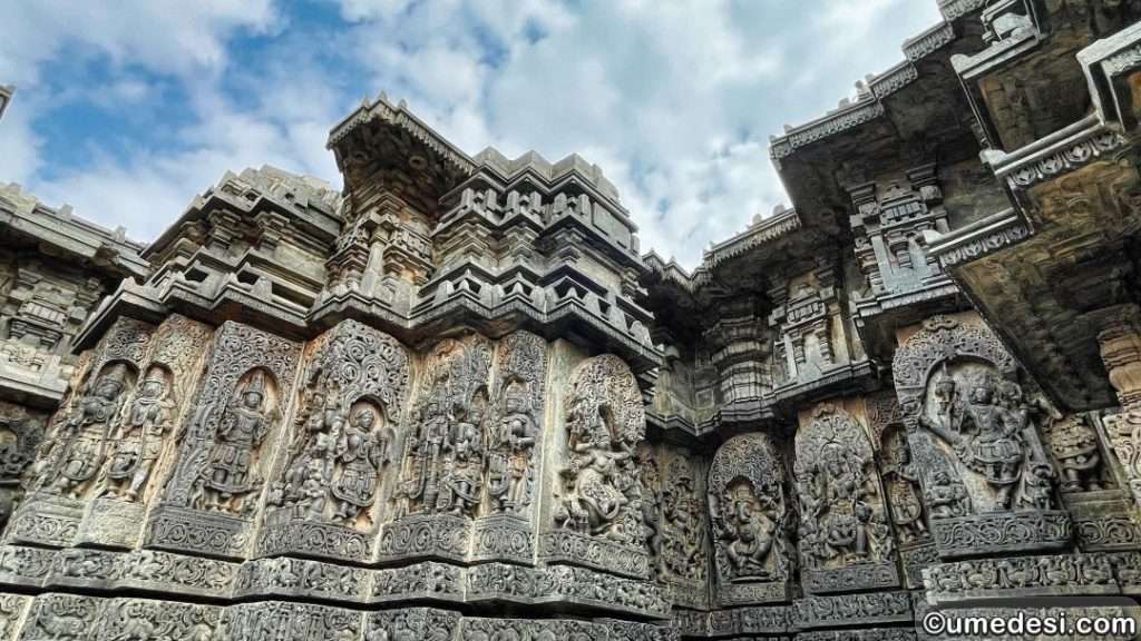 A wide-angle view of the exterior of Hoysaleswara Temple, adorned with intricate wall carvings under a blue sky.