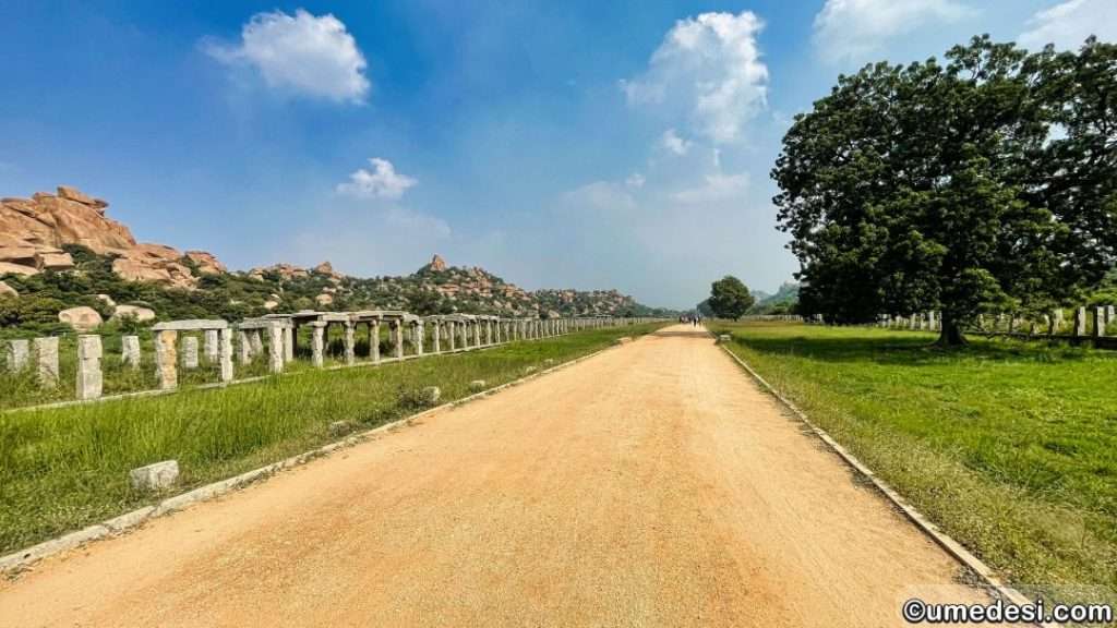 Pathway between remnants of ancient store pillars amidst greenery in Hampi's Vijaya Vittala temple complex.